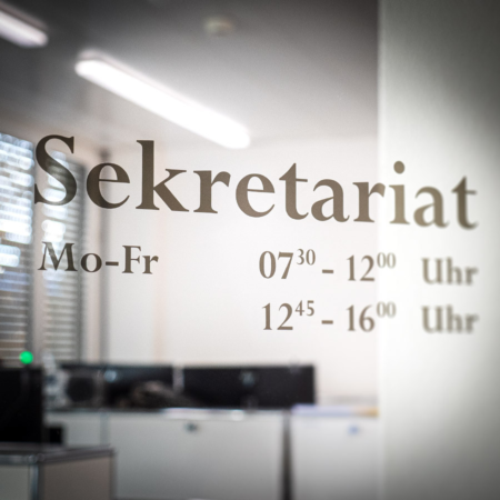 Beschriftung "Sekretariat inkl. Öffnungszeiten" an Glaswand.