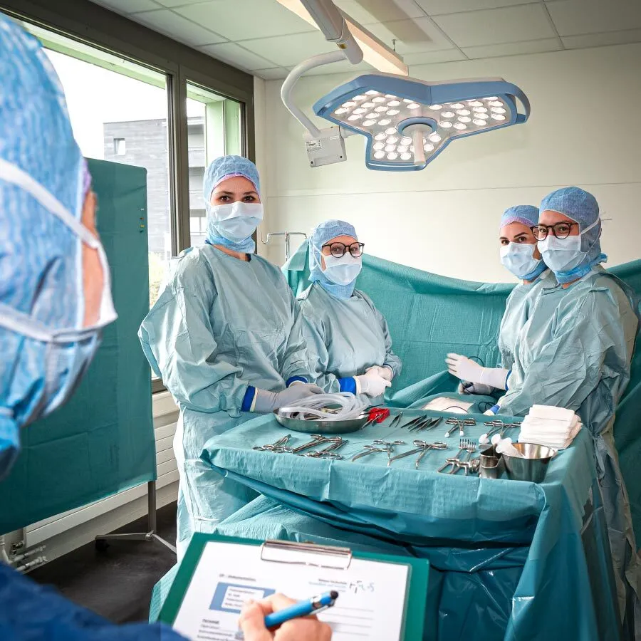 Vier Personen in Operationskleidung stehen um einen Tisch mit Operationsbesteck und schauen zu einer fünten Person im Bildvordergrund