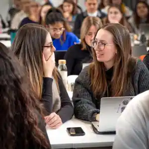 Zwei Studentinnen sitzen im Hörsaal der HFGS zusammen mit anderen Studierenden vor dem Laptop und reden miteinander